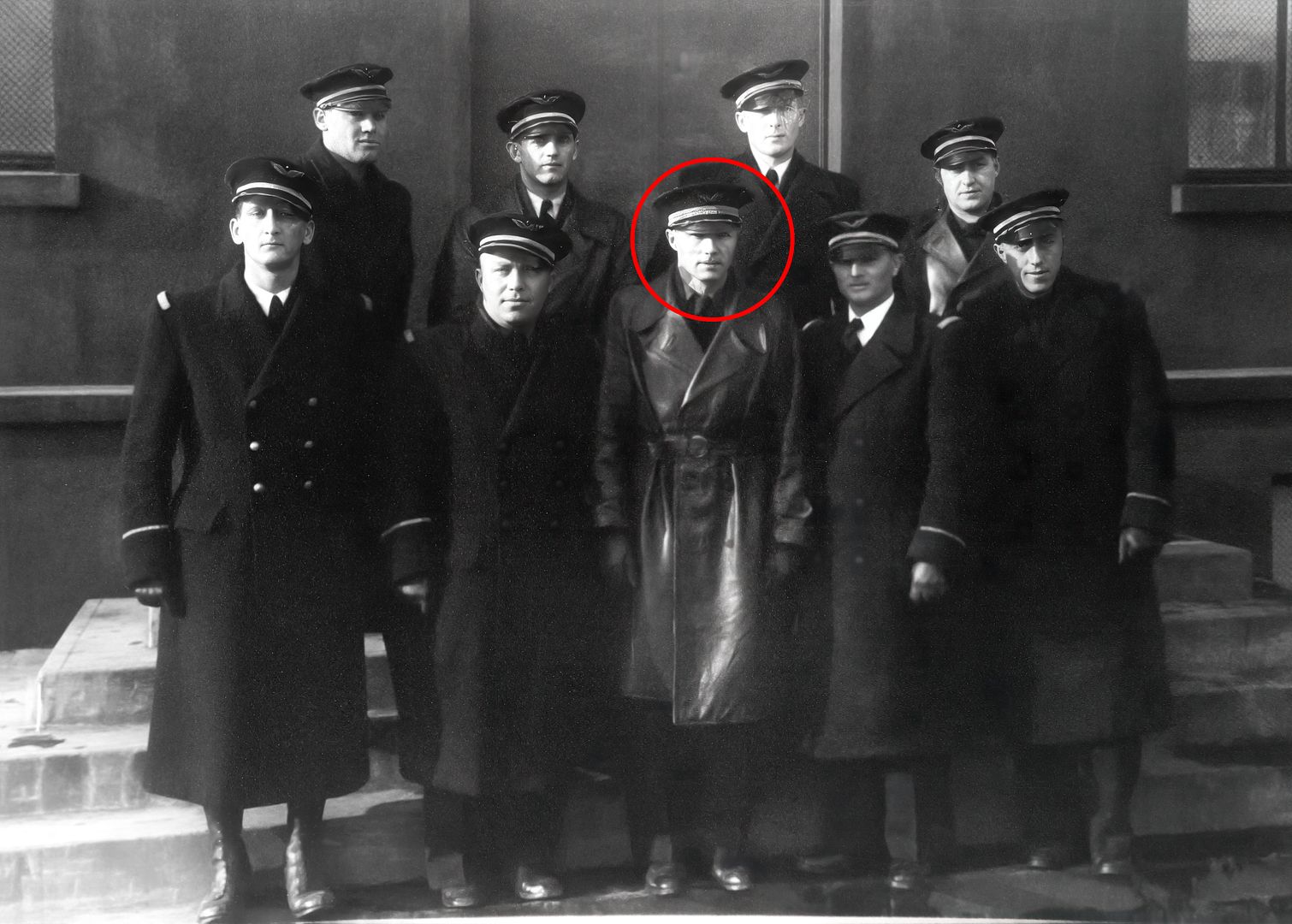 A vintage black and white photograph of nine men, all dressed in military-style uniforms and hats, standing together in a semi-formal pose. The central figure, highlighted with a red circle, wears a long leather coat and a distinctive hat.
