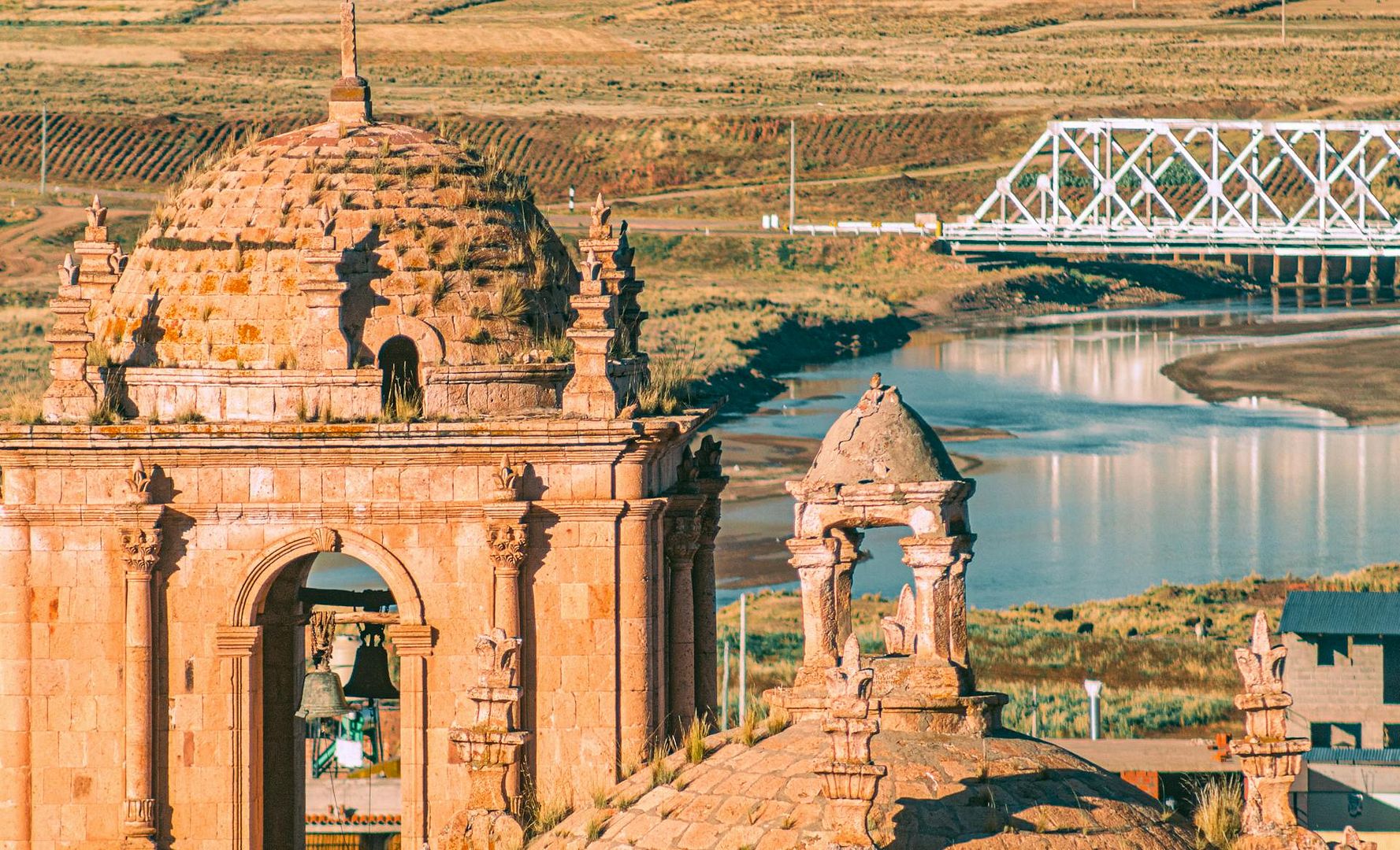 Close-up of a colonial stone bell tower and domes overlooking a river and a white bridge in a rural Andean valley in Puno, Peru