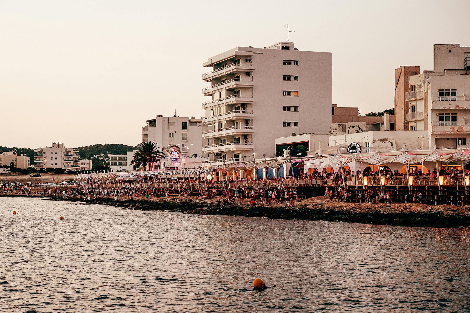 Crowded beach clubs and white hotels along the rocky Ibiza coast at sunset, featuring Café Mambo.