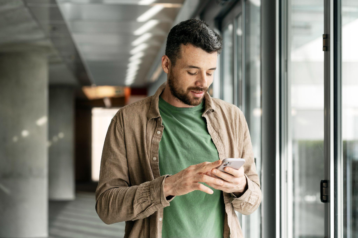 A man standing indoors, focused on reading a message on his mobile phone.