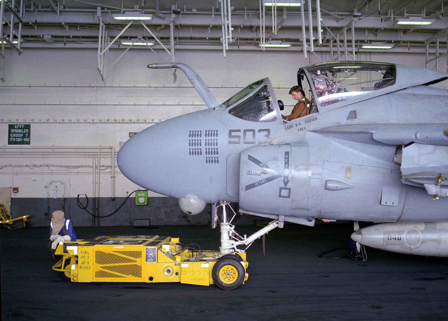 A Crewman Manning An SD 1D Aircraft Spotting Dolly Waits While A Shipmate Inspects The Cockpit Of An Attack Squadron 75 A 6E Intruder Aircraft N The Hangar Deck Of The Aircraft Carrier USS KITTY HAWK