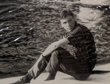B&W George in front of body of water in 50s. A black and white photo shows a white man sitting in front of a body of water. He is looking to the side with a serious expression and has short, dark hair and a cleft chin. He is wearing a short-sleeved, collared, plaid top, a dark wristwatch, and dark slacks.