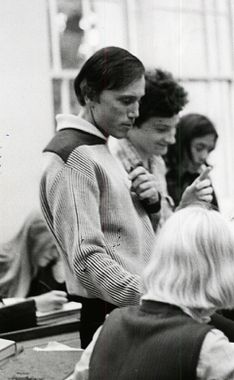 Bergman, George '76 at GFS. A black and white photo focused on a dark-haired white man with hair to his ears wearing a striped, collared sweater. He is looking down and one hand is visible holding something casually in front of his chest. There are blurry other people around him—one in the foreground with chin-length blond hair, and one right behind him with dark curly hair.
