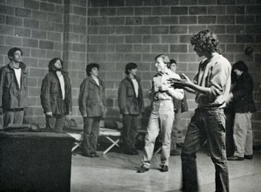 George Bergman in The Brig at GFS in 1977. A black and white photo shows people on a stage with a brick background. Towards the back, five men are lined up in front of cots as if at attention. They are wearing white shirts with dark, loose-fitting jackets and dark pants. A thin white man with light, ear-length hair stands in the middle wearing a light collared top and light slacks. His elbows are at his sides and his hands touch over his ribs while he faces the people who are lined up. Another man in the foreground holds his hands up as if explaining something. His hair is brown and curly and he's wearing flared jeans and a light sweater.