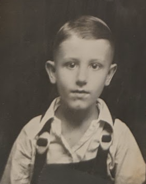 George as a child in the 1930s. A black and white photo shows a young boy in a collared white shirt and dark overalls looking at the camera. He has short, dark hair slicked back and to the side, dark eyes, prominent ears, and a dimple in his chin. The background is black.