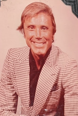 George in houndstooth in the 70s or 80s. A tan white man with light, ear-length hair, dark eyes, and a cleft chin smiles open-lipped at the camera against a pink background. He is wearing a brown and white houndstooth sport coat over a dark top.