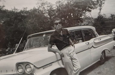George leaning against car in the 50s. A black and white photo with bushes in the background shows a long white car, probably from the 50s, in the foreground. A tan white man with short, dark hair leans back with his elbow on the car while looking at the camera. He has a cleft chin and wears large, dark sunglasses, a dark, short-sleeved, collared, striped top, and light slacks.