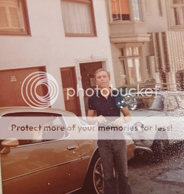 George possibly in San Francosc. A thin white man with short, light hair, a cleft chin, and a prominent Adam's apple looks toward the camera with his chin up. He is wearing a dark polo shirt, a dark wristwatch, grayish-brown flare pants, and a thick dark belt with a silver buckle. Behind him are cars, maybe from the 70s—one brown and one off-white. Behind the cars are buildings possibly in San Francisco — one with a flat, white front and reddish garage, door, and window frames, and a beige one with a bay window.