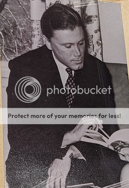 George in suit, pointing with pen. A black and white photo shows a white man with a strong cleft chin and his short, dark hair combed back. He is wearing a dark suit and tie and sitting with one hand on his knee and his other hand holding a pen to point at something in a book that someone cropped out of the picture is holding.