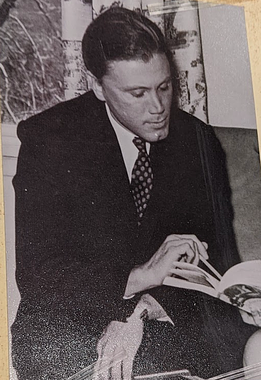 George in suit, pointing with pen. A black and white photo shows a white man with a strong cleft chin and his short, dark hair combed back. He is wearing a dark suit and tie and sitting with one hand on his knee and his other hand holding a pen to point at something in a book that someone cropped out of the picture is holding.