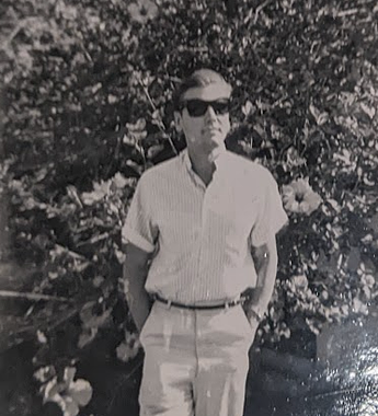George in sunglasses. A black and white photo shows a white man with short, dark hair combed back. He is standing in front of a large, flowered bush with his hands in his pockets. He is wearing large, dark sunglasses, a short-sleeved, collared, striped, light top with the sleeves rolled up, and light pants with a thin dark belt.