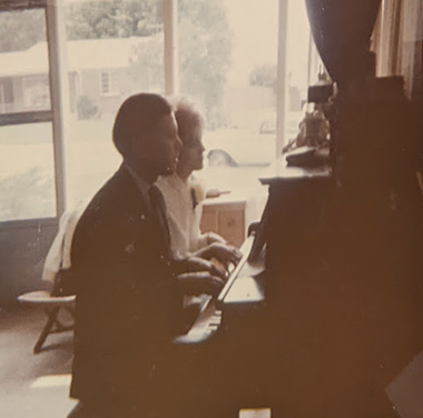 Carroll and George at the piano. A white man and white woman are backlit as they play a duet at a dark, upright piano. He has short, dark hair and is wearing a suit and tie. She has light hair and is wearing a white collared top with a flower on one side.