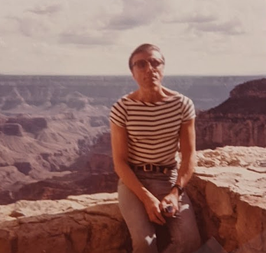 George at Grand Canyon. A thin white man looking at the camera with a serious expression sits on a rock wall at what appears to be the Grand Canyon. He has short, dark hair, a striped boat neck top, a dark belt with a silver buckle, a black wristwatch, form-fitting jeans, and he is wearing large, translucent sunglasses.