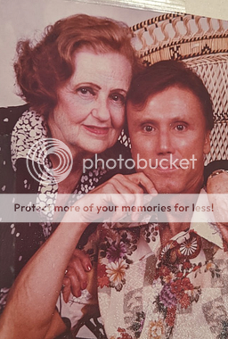 George and his mother. On the left is a white woman with reddish, ear-length hair, red lipstick, drawn-in brown eyebrows, and a black and white collared top. She has one arm around a dark-haired white man on the right, with her hand on his shoulder, her other hand on his bicep, and her head touching his. On the right, the man is seated in a macramé-like chair and leans his elbow on the arm of the chair, with his hand up to his dimpled chin. He has short, dark hair and is wearing a  floral and geometric collared top. Both are looking at the camera with a pleased look.