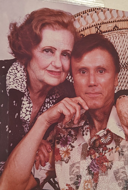 George and his mother. On the left is a white woman with reddish, ear-length hair, red lipstick, drawn-in brown eyebrows, and a black and white collared top. She has one arm around a dark-haired white man on the right, with her hand on his shoulder, her other hand on his bicep, and her head touching his. On the right, the man is seated in a macramé-like chair and leans his elbow on the arm of the chair, with his hand up to his dimpled chin. He has short, dark hair and is wearing a  floral and geometric collared top. Both are looking at the camera with a pleased look.